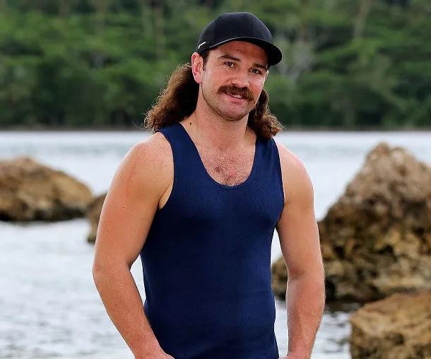Man in a blue tank top and cap standing near rocks with water and trees in the background.