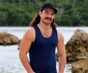 Man in a blue tank top and cap standing near rocks with water and trees in the background.