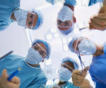 Surgeons in blue scrubs and masks, seen from below, performing an operation under bright surgical lights.