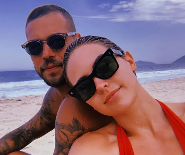 Couple with sunglasses on a beach, ocean waves and mountains in the background, under a partly cloudy sky.