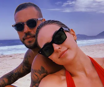 Couple with sunglasses on a beach, ocean waves and mountains in the background, under a partly cloudy sky.
