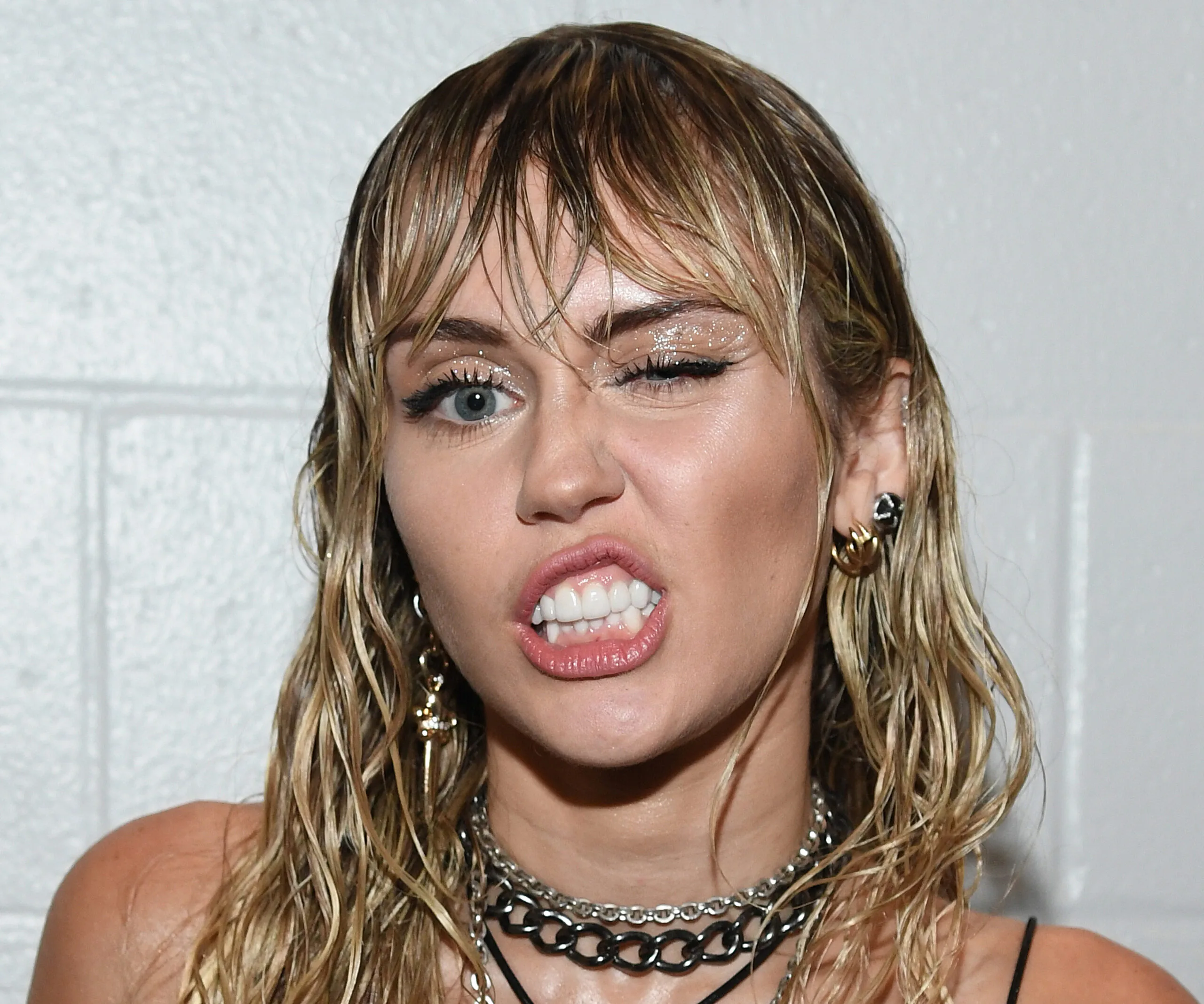 Singer with wet hair winking, showcasing chain necklaces and bold earrings in a playful expression.