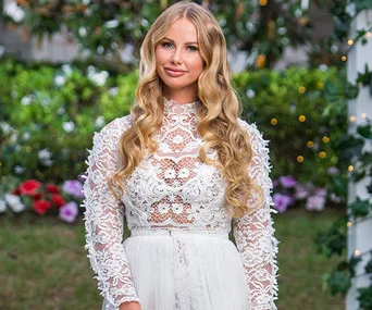 A woman in a detailed white lace dress stands outdoors, with flowers and greenery in the background.