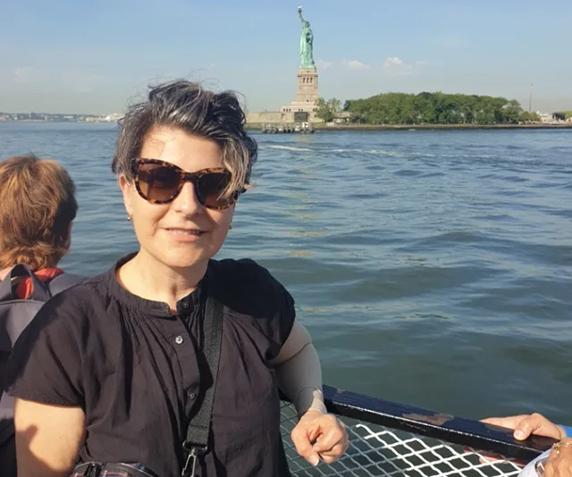 Woman on boat with sunglasses smiles at camera, Statue of Liberty visible in background.