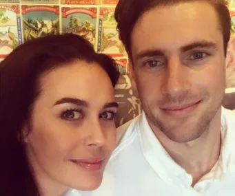 Two people pose closely together, smiling softly in front of a colorful, patterned background.