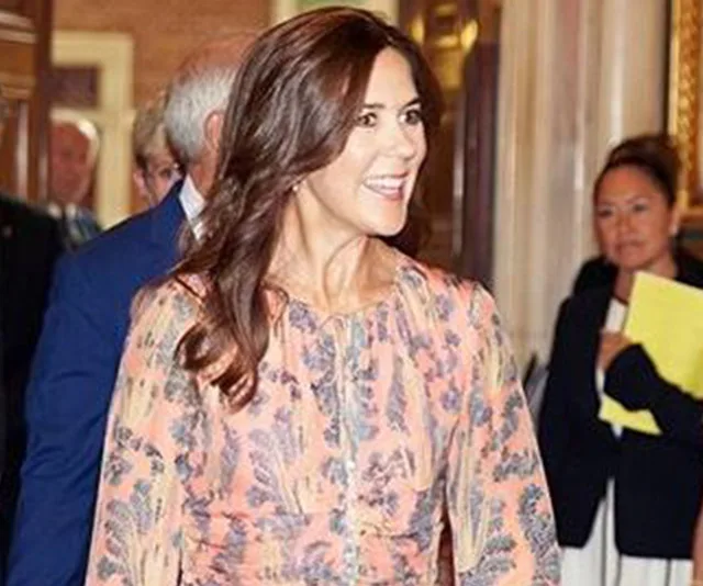 A woman in a floral dress smiles while walking indoors, with others in the background.