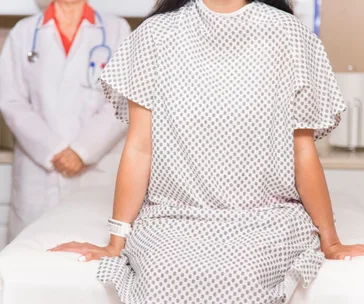 "Patient in a patterned hospital gown sits on examination table, with a doctor standing in the background."