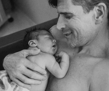 A man smiles while holding a sleeping newborn baby close to his chest, capturing a tender and intimate moment.