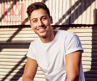 Man in a white t-shirt smiles in front of a corrugated metal wall with "TV WEEK" logo visible.