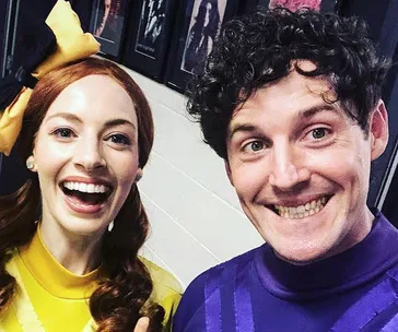 Two people smiling; one in a yellow outfit with a bow, the other in purple. Photos on the wall in the background.