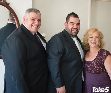 Three people smiling in formal attire; two men in suits and a woman in a purple dress, with a "Take5" logo visible.