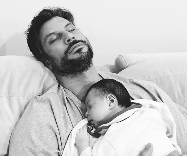 Man sleeping on a couch with a baby resting on his chest.