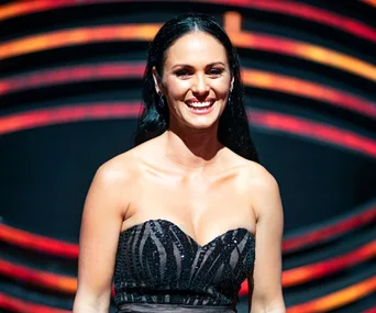 Woman in a strapless black dress smiling on stage with a vibrant circular background.
