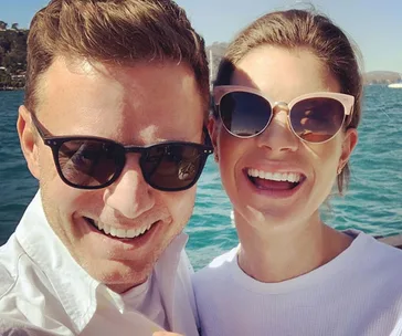 A couple wearing sunglasses smiles outdoors with a sunny waterfront background.