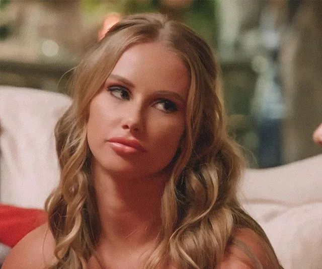 A woman with long wavy hair looks sideways with a serious expression, seated indoors.