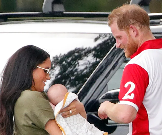 Mother holding baby while standing next to a man in a red-and-white polo shirt, outdoors beside a car.