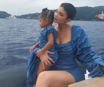Mother and child in matching blue outfits sit on a boat, with a scenic ocean backdrop and distant yacht.