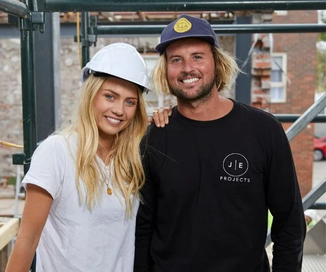 A couple on a construction site; one wearing a hard hat and the other in a cap and black shirt, smiling at the camera.