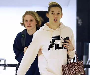 A woman in a FILA hoodie holding a phone, accompanied by a man with a backpack, walking in an airport.
