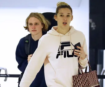 A woman in a FILA hoodie holding a phone, accompanied by a man with a backpack, walking in an airport.