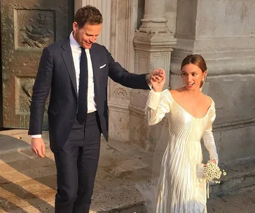 A newlywed couple exits a church, with the groom in a navy suit and the bride in an elegant white gown holding a bouquet.