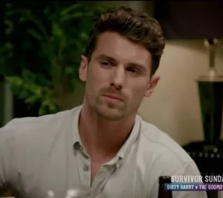 A man sitting indoors, wearing a light button-up shirt, looking to the side with a thoughtful expression.