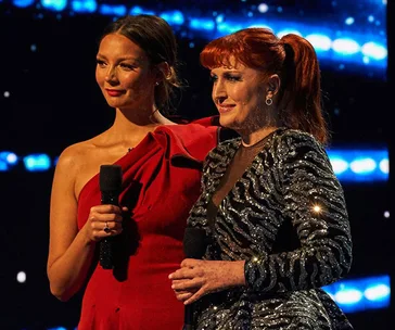 Two women stand on stage holding microphones, wearing a red dress and a sparkly black dress, with a blue-lit background.