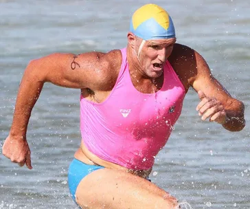 Athlete in a pink swimsuit with a yellow-blue cap runs through shallow water, muscles tense and water splashing.