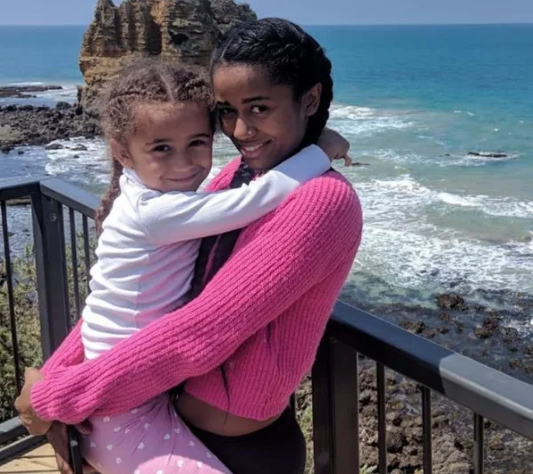 Mother and daughter sharing a hug on a coastal boardwalk, with a scenic ocean backdrop.