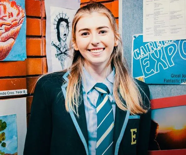 Georgie Stone as Mackenzie Hargreaves in school uniform, smiling in front of a bulletin board with student artwork.