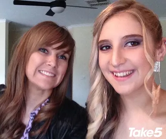Two women smiling in a room, one with straight brown hair and the other with curled blonde hair, wearing earrings.