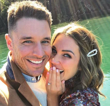 A couple smiling by a lake, with the woman showing an engagement ring and covering her mouth in surprise.