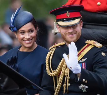 Prince Harry and Meghan Markle in formal attire, smiling and waving during a carriage procession.