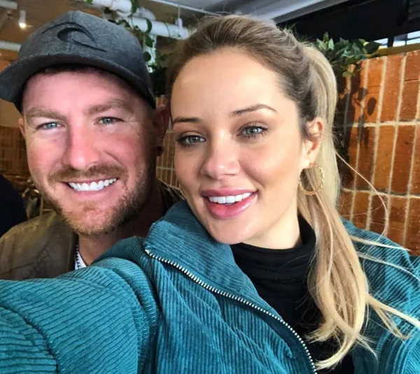 Two people smiling for a selfie indoors, one wearing a cap and the other in a teal jacket with gold hoop earrings.