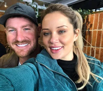 Two people smiling for a selfie indoors, one wearing a cap and the other in a teal jacket with gold hoop earrings.