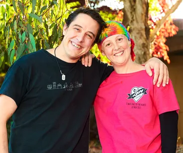 A smiling man in a black shirt and a woman in a pink "Love Your Sister" shirt stand together outdoors.