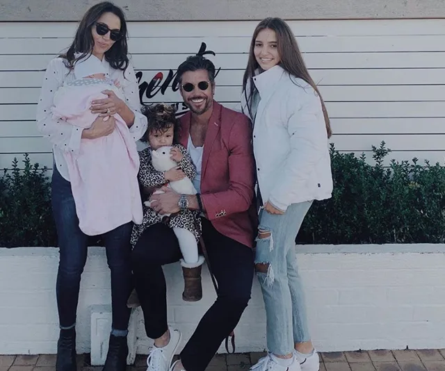 Family photo with two adults and two children posing outdoors, one child held by the man, and a baby with the woman.