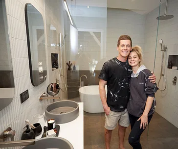 Couple posing in modern bathroom with grey sinks, mirrors, and a freestanding tub in the background.