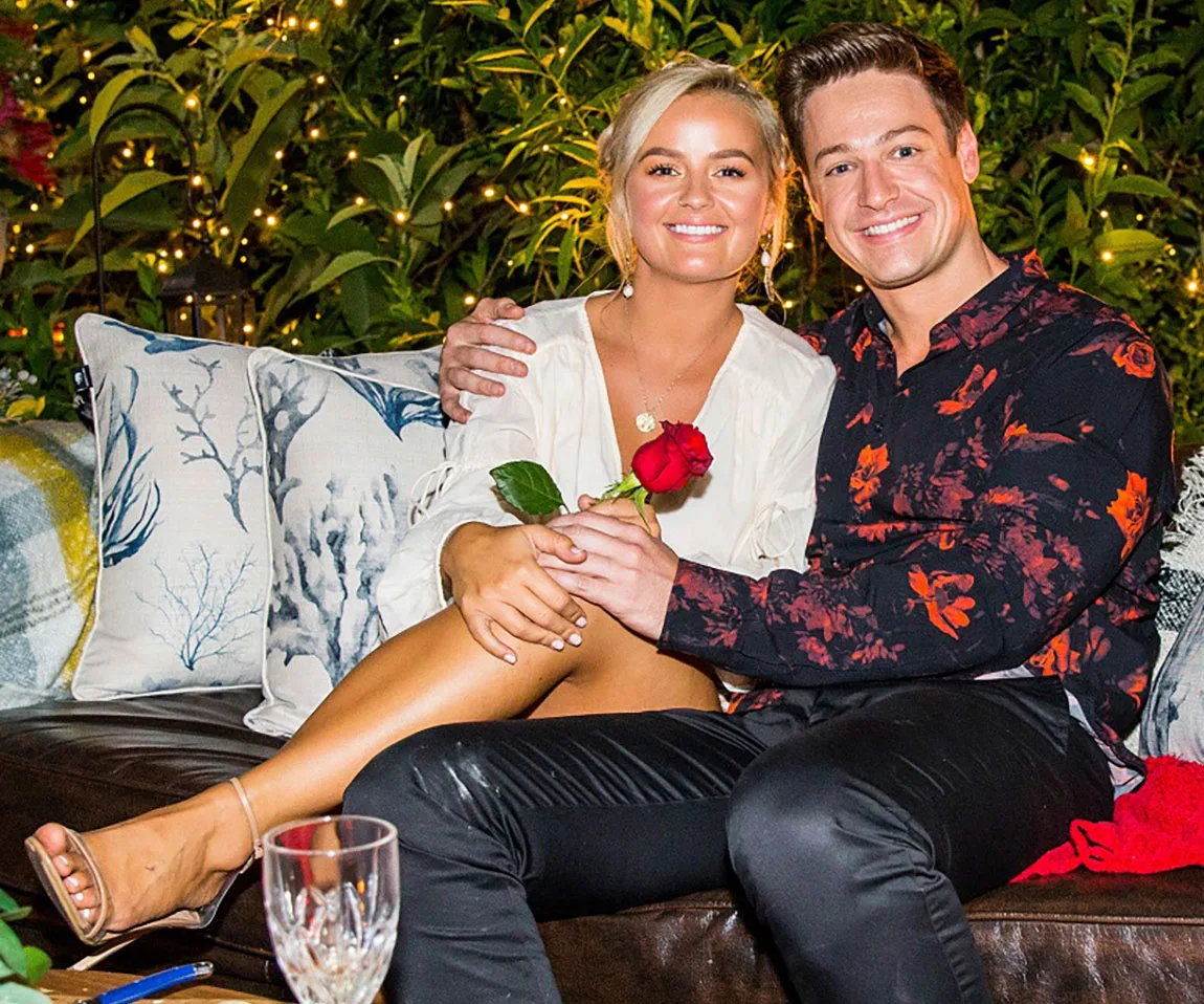Couple smiling and sitting on a sofa outdoors, holding a rose, surrounded by decorative pillows and plants.