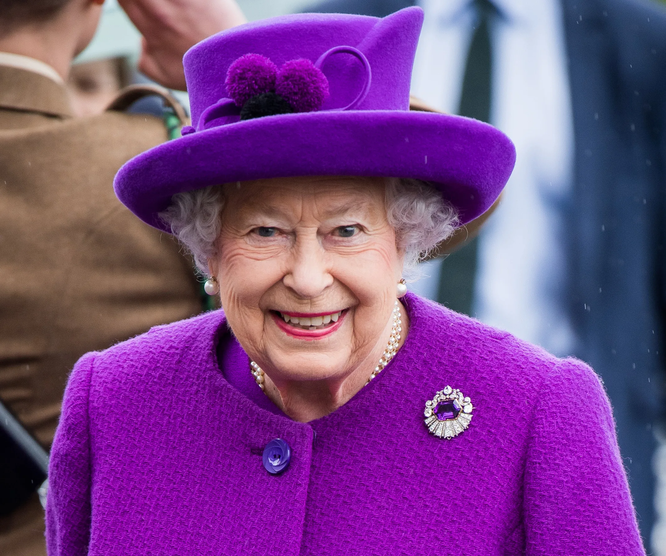 A person smiling in a vibrant purple outfit and hat, adorned with a brooch, during an outdoor event.
