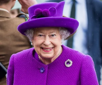 A person smiling in a vibrant purple outfit and hat, adorned with a brooch, during an outdoor event.