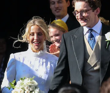 Ellie Goulding in a long-sleeved white wedding dress and veil, holding flowers beside her groom, smiling.