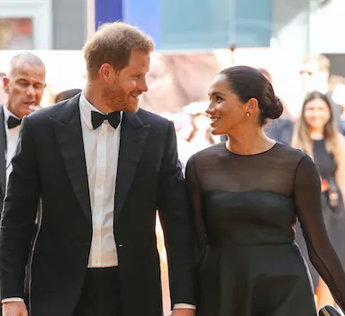 Prince Harry and Meghan Markle smiling at each other on a red carpet.