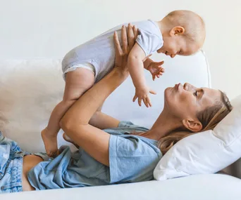 A mother joyfully lifts her smiling baby in the air while lying on a white couch.