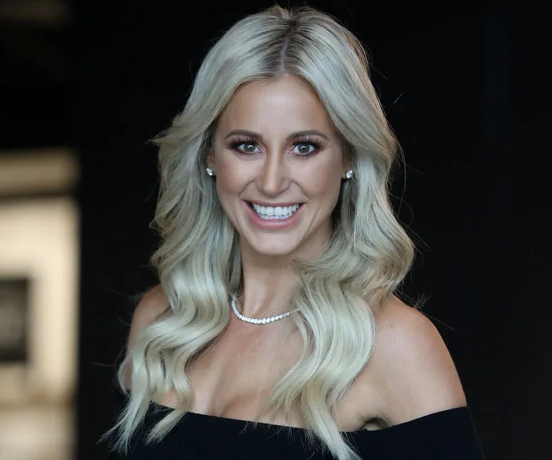 A woman with long blonde hair smiling, wearing an off-shoulder black top and pearl necklace.