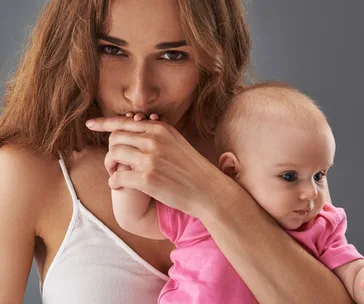 Mother affectionately kissing baby's hand while holding her. Baby is in a pink outfit.