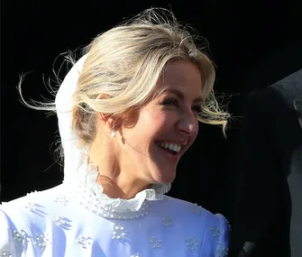 A bride in a white gown with floral details and a high collar, smiling beside a person in dark attire.