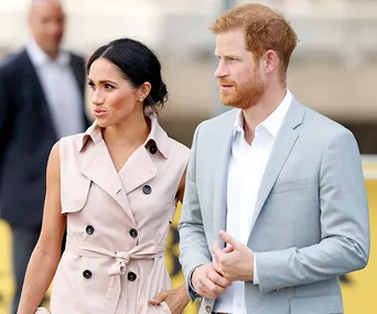 A woman in a sleeveless trench coat and a man in a light suit walking outdoors, looking in the same direction.