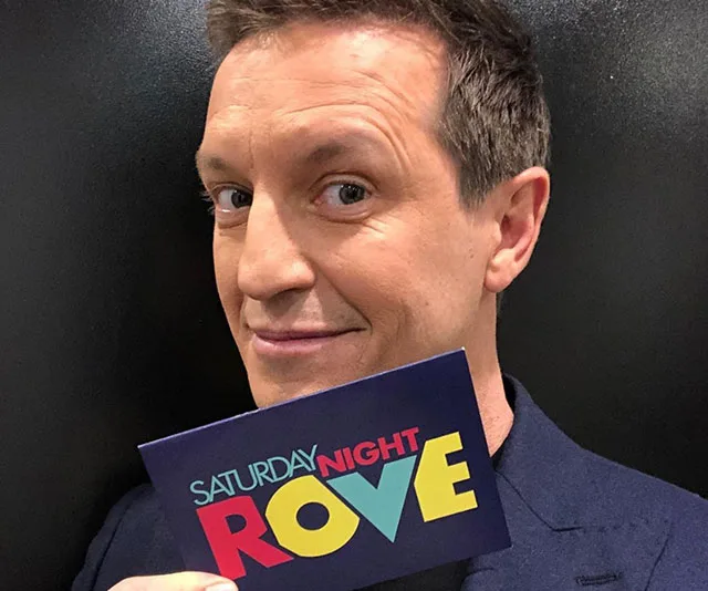 Man holding "Saturday Night Rove" card, smiling at camera.