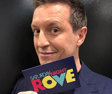 Man holding "Saturday Night Rove" card, smiling at camera.